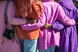 In a view of their backs, four women hug each other standing in a line. All wear bright coloured clothes.