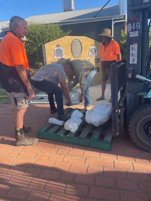 Dois homens levantam um grande pedaço de uma estátua quebrada e o colocam em uma empilhadeira enquanto outro homem observa.