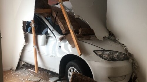 The nose of a car protruding from a smashed wall inside a house.