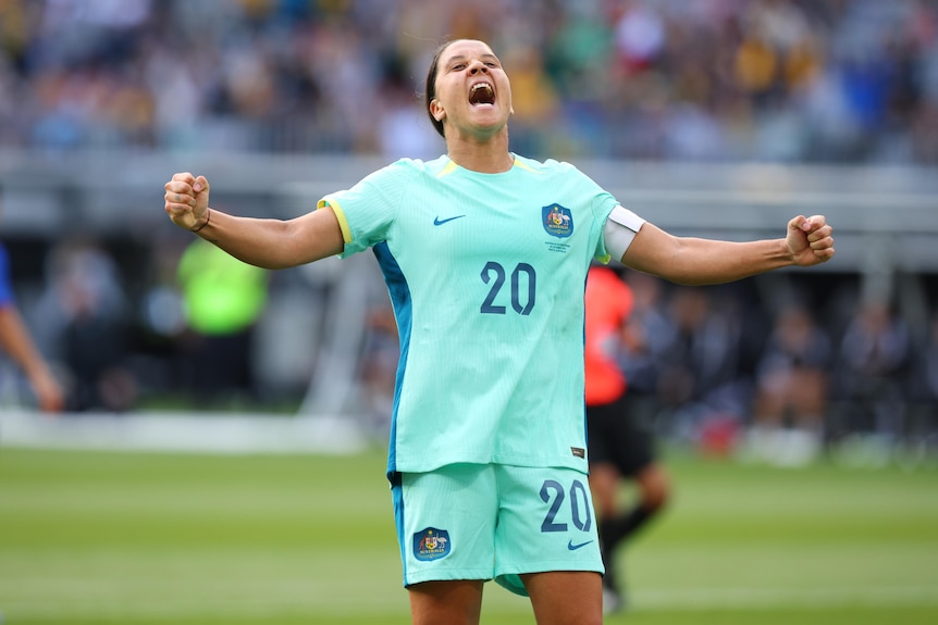 A soccer player wearing light blue holds her arms out and yells during a game