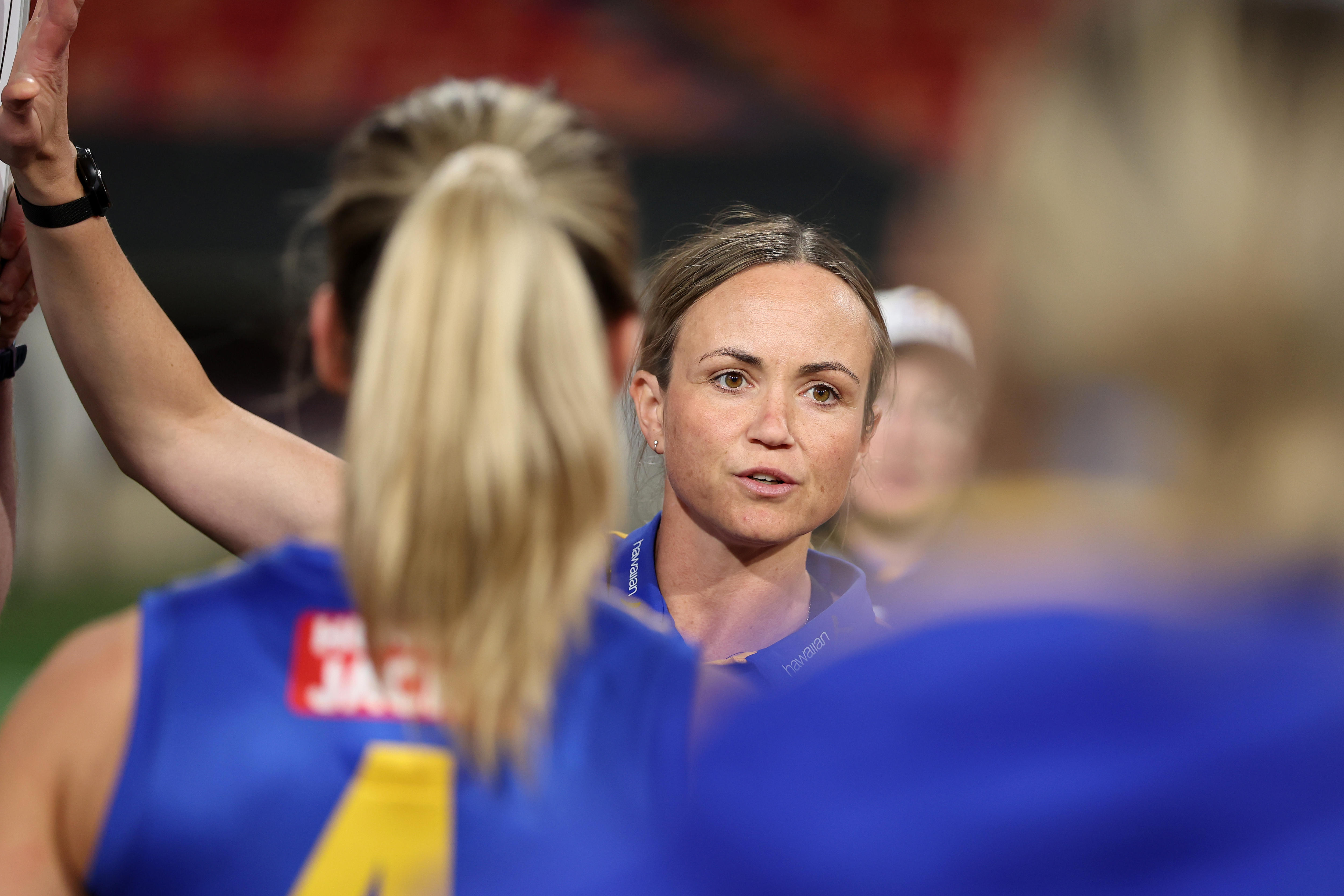 A West Coast AFLW coach looks out at her players (who have their backs to the camera) during a team talk in a game.