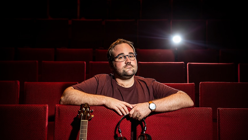 A white middle-aged man in a theatre, with a guitar and headphones, frowning