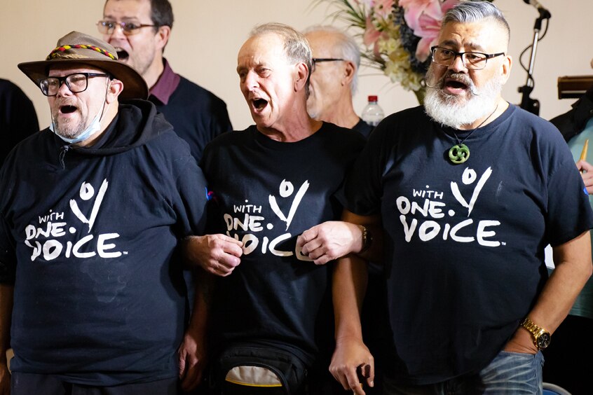 Three men with linked arms open their mouths to sing