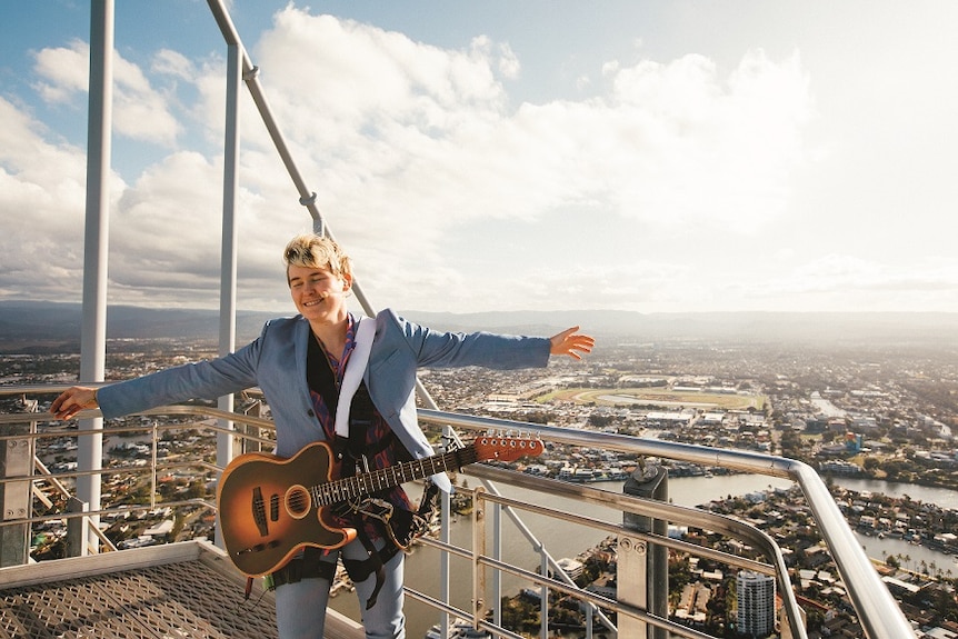 A musician with a guitar shuts her eyes and holds out her arms on top of the very tall Q1 building on the Gold Coast.