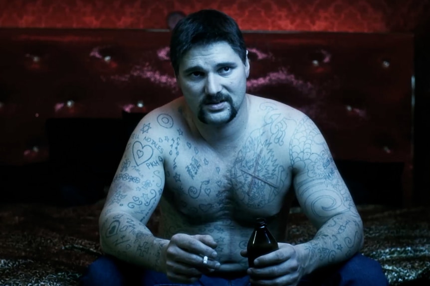 Beefy 30-something man with handlebar mustache sits bare-chested on leather couch, torso covered in tattoos.