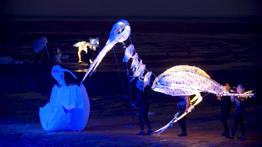 A theatre performance on a beach showing delicately constructed large puppets operated by people with long poles.