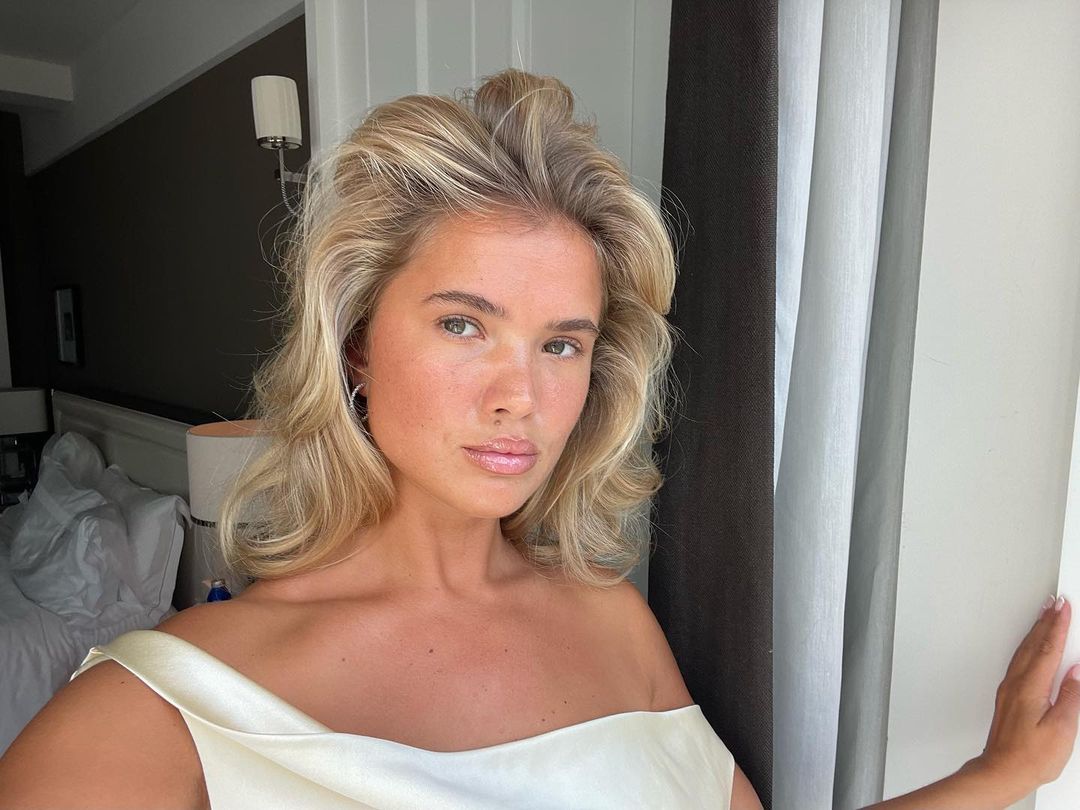 A tanned woman with blonde voluminous hair looks at the camera. 