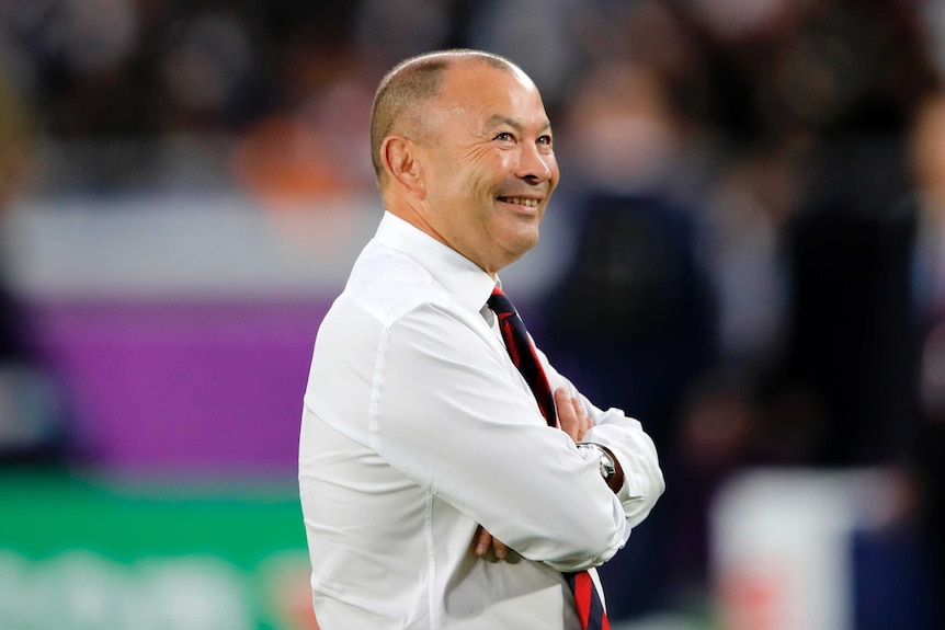 Eddie Jones, wearing a shirt and tie, smiles widely while looking up towards the stands in the stadium.