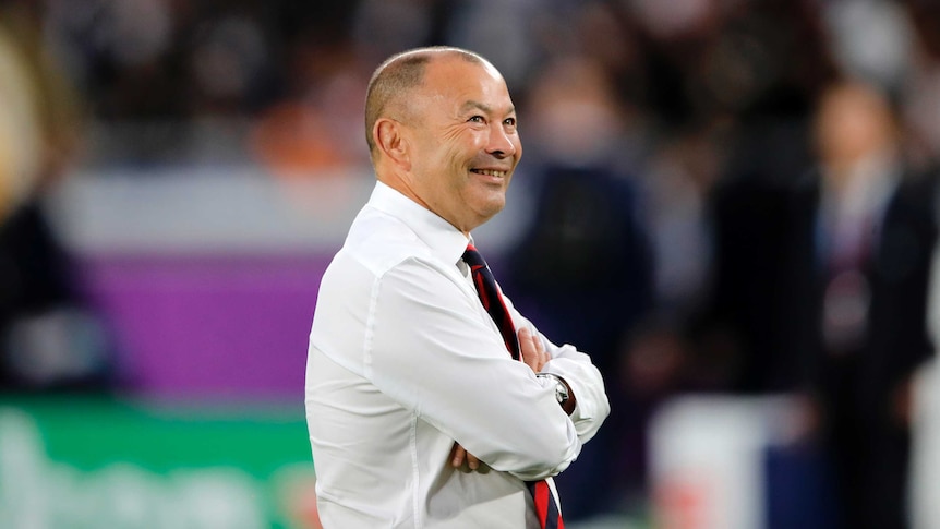 Eddie Jones, wearing a shirt and tie, smiles widely while looking up towards the stands in the stadium.