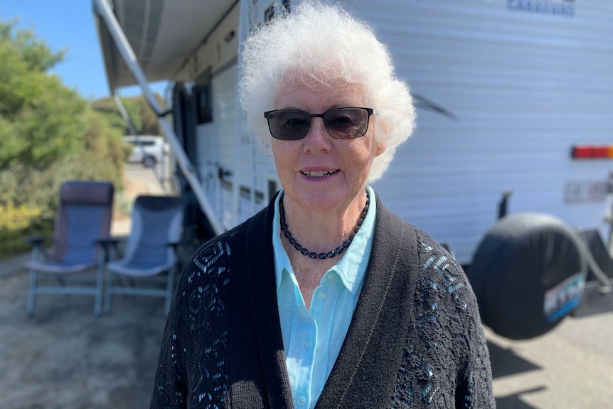 Marilyn Halladay, wearing a cardigan and sunglasses, smiles at the camera with a caravan in the background.