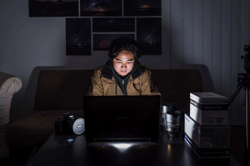 Paean Ng sits in a darkened room with his face illuminated by a laptop computer.