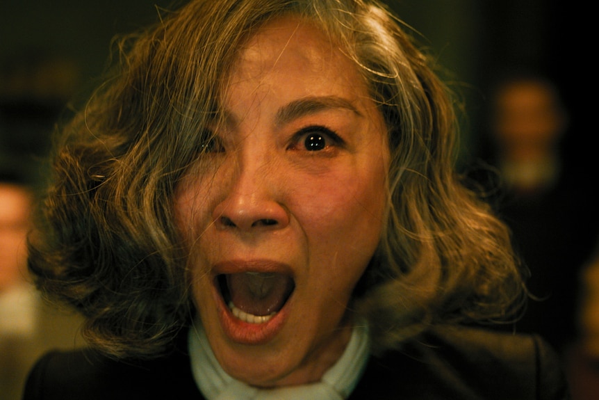 A close up of a woman with grey hair, eyes wide with terror and mouth open, perhaps screaming