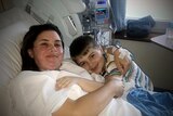 A woman in a hospital bed with a child at her bedside hugging her and smiling. 