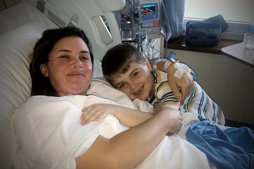 A woman in a hospital bed with a child at her bedside hugging her and smiling. 