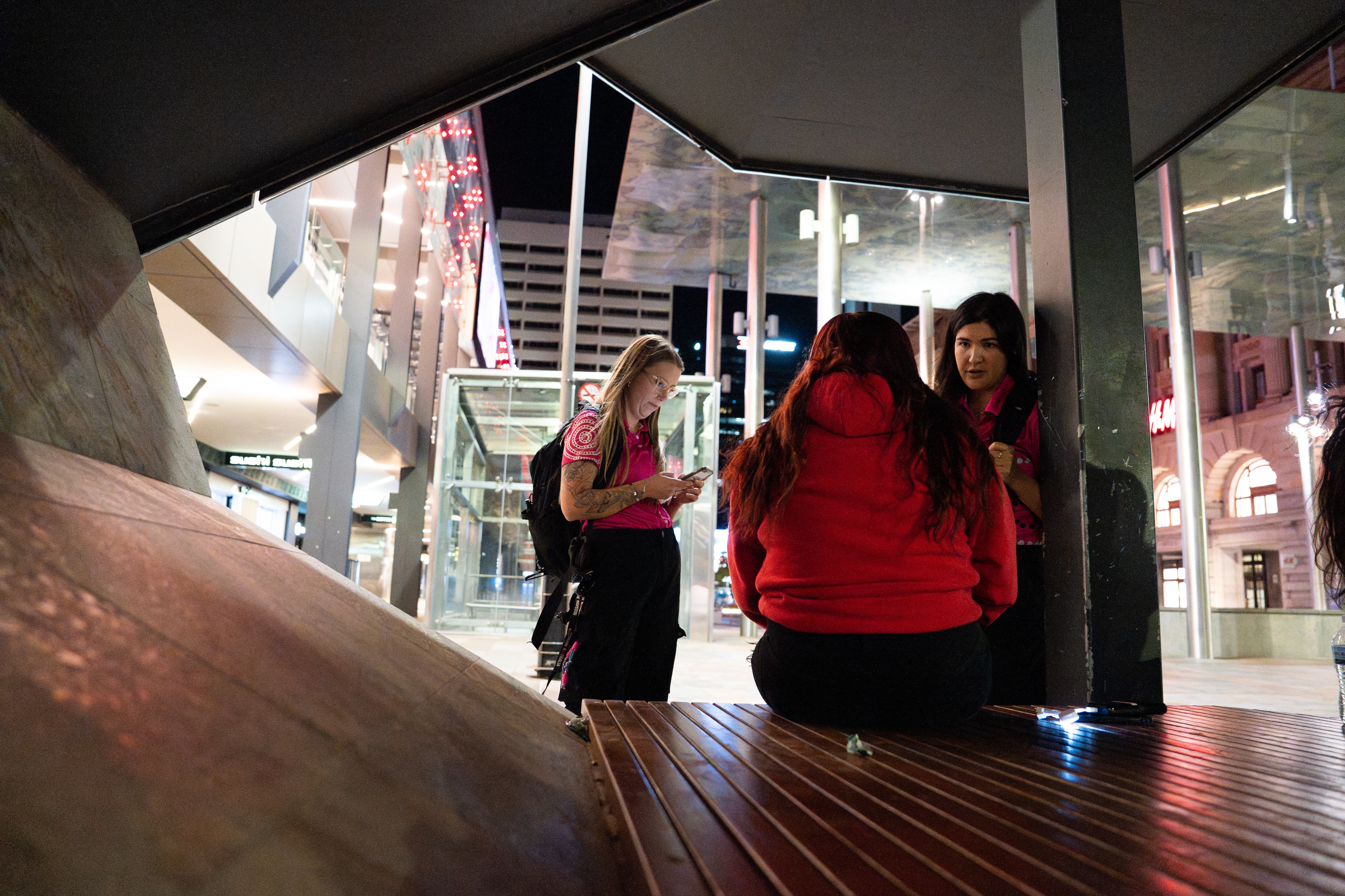 Two women who are youth workers stand in a Perth street at night talking to a teenage girl in a red jumper.