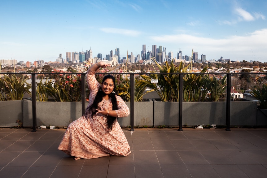 You view a woman of South Asian descent pictured in a traditional Indian dress and classical dance pose on a rooftop.