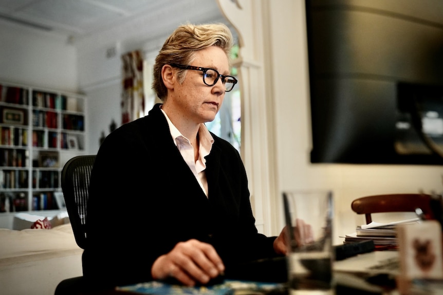 A middle-aged blonde woman with short hair sits at her computer with her right hand on a mouse.