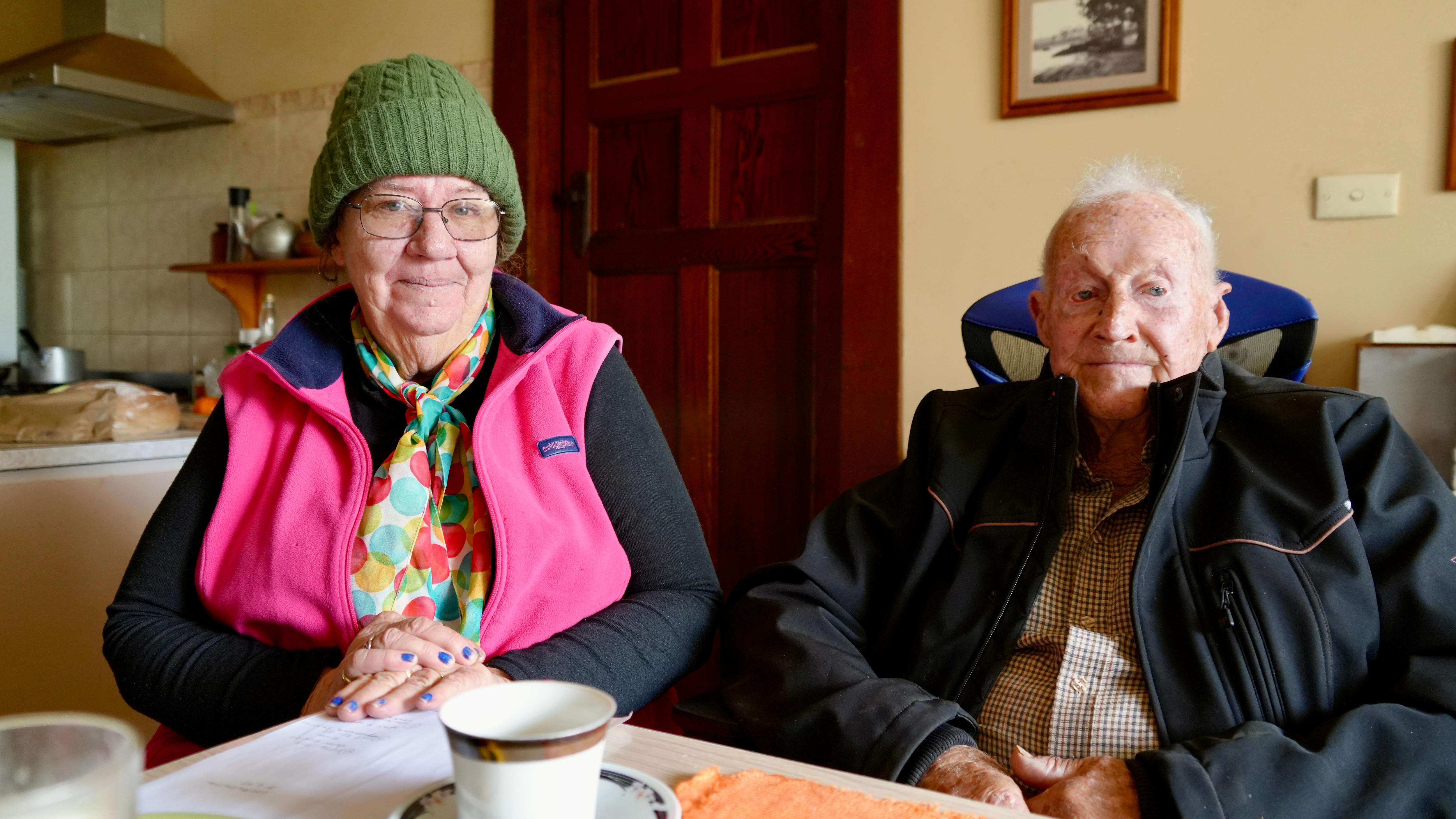 a lady in a pink vest and beanie with glasses and a older man with grey hair and a black jacket smiling at the camera 