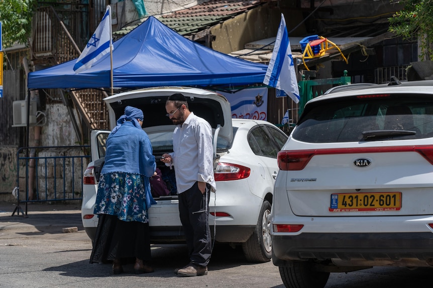 A Jewish couple talk next to a car with Israeli flags erected behind them 