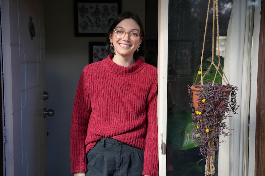 A woman wearing a red sweater smiles while standing in her front doorway.