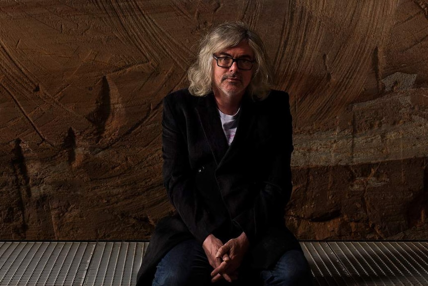 Man with grey shoulder-length hair and black-rimmed glasses in black suit kneeling on metal grate with sandstone wall behind.