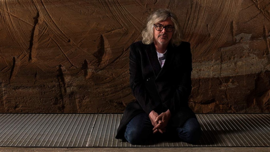 Man with grey shoulder-length hair and black-rimmed glasses in black suit kneeling on metal grate with sandstone wall behind.