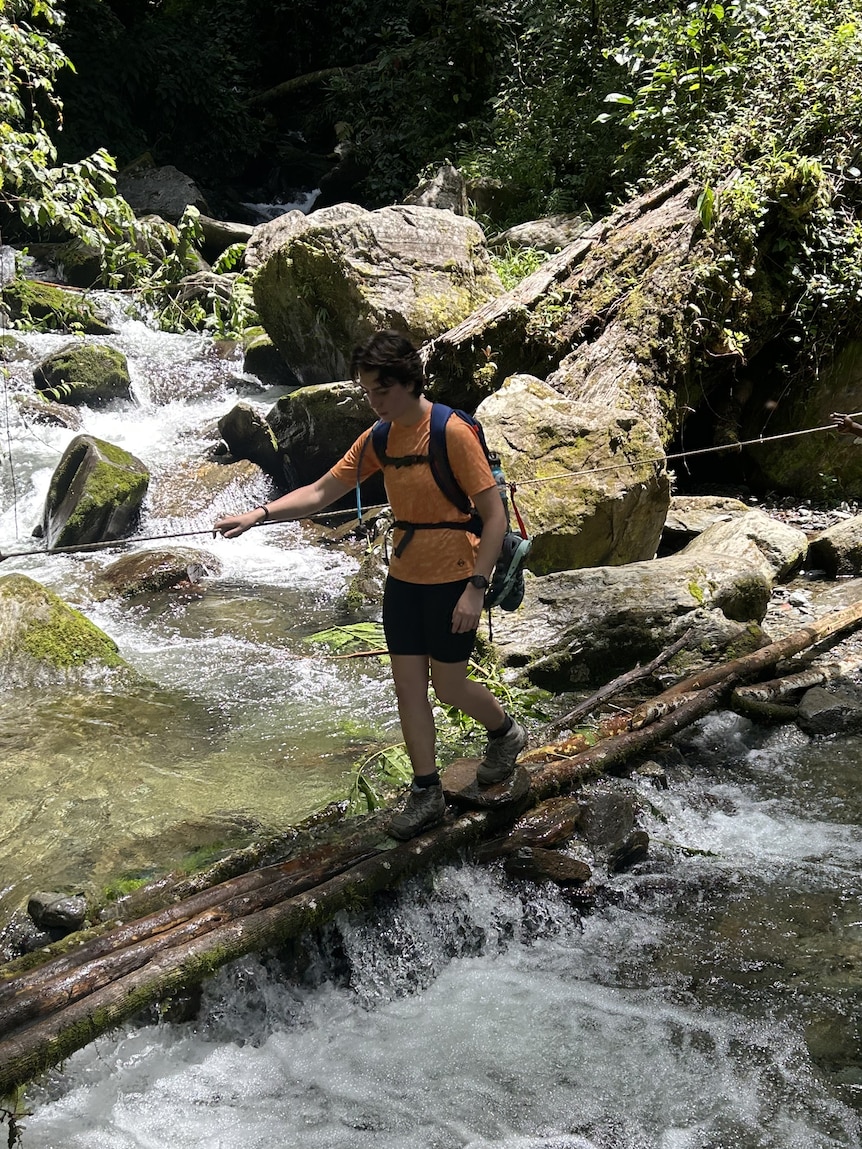 A young woman wearing a backpack crosses a river by walking over a branch. 