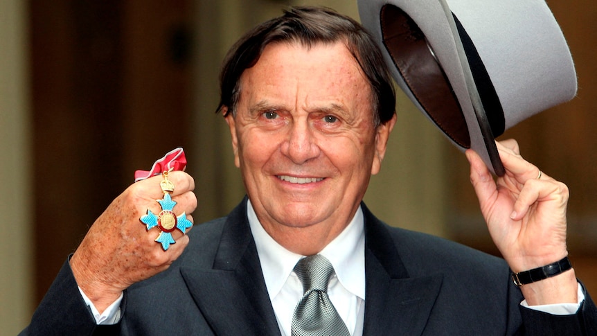 Barry Humphries tips his hat as he holds up a medal. 