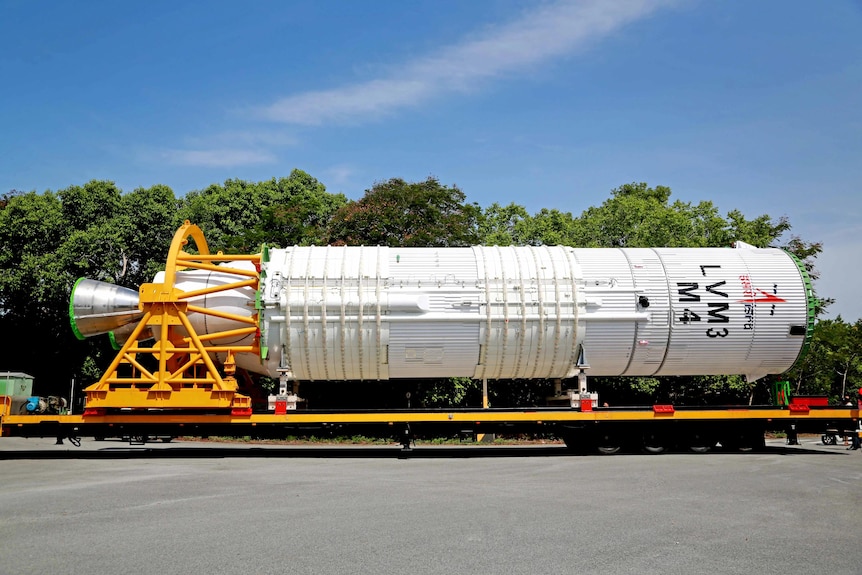 A large space rocket lies on the yellow bed of a truck as it is moved along a road.