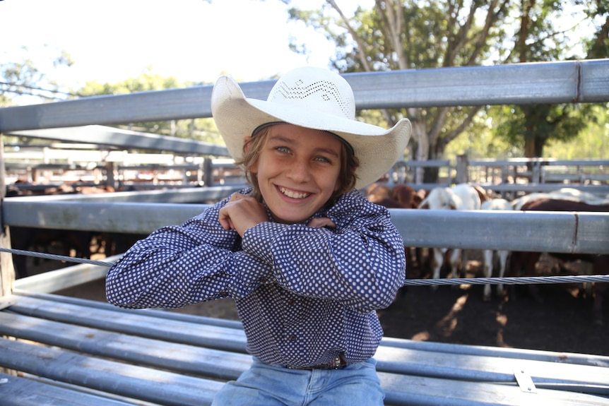 A young boy in an cowboy hat leans on a fence