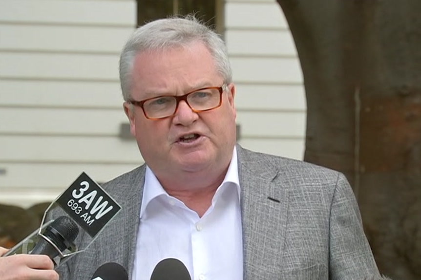 A man with glasses in a suit holds a media conference under a tree.