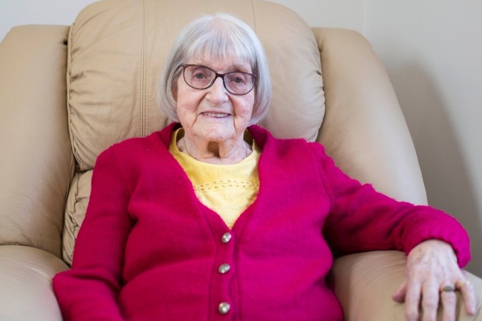 An elderly woman in a red cardigan sits smiling in a lounge chair