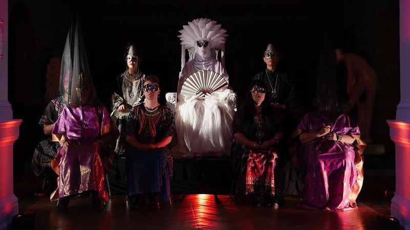 Performers dressed in elaborate costumes sit on a stage.