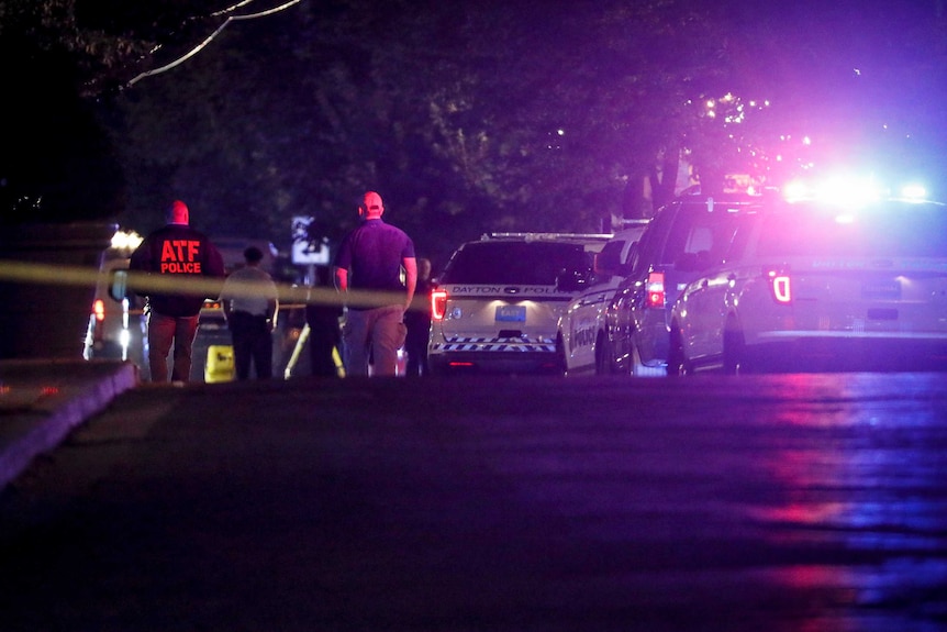 Four police cars are seen in the glow of blue light, with four people seen to the left walking along a road in darkness.