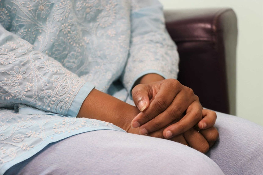 A woman sits on a couch with her hands clasped in her lap.