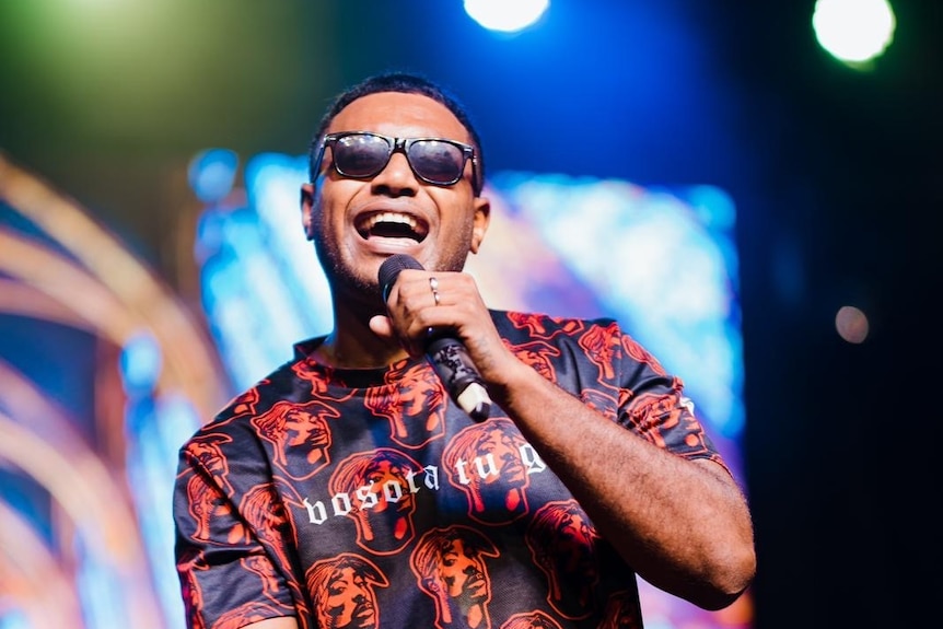 A man wearing sunglasses holds a microphone, performing onstage.