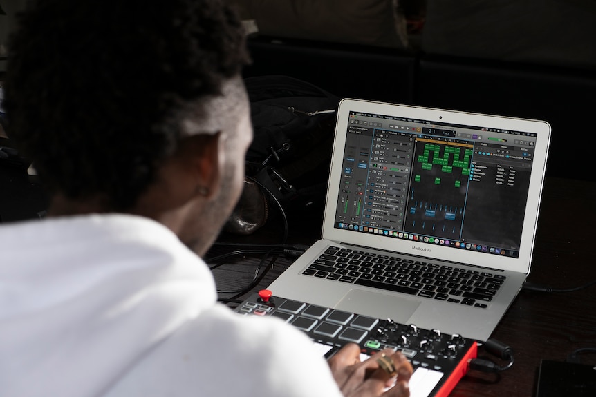 Looking over the shoulder of Egrinya Adie while he produces beats using a laptop and MIDI keyboard