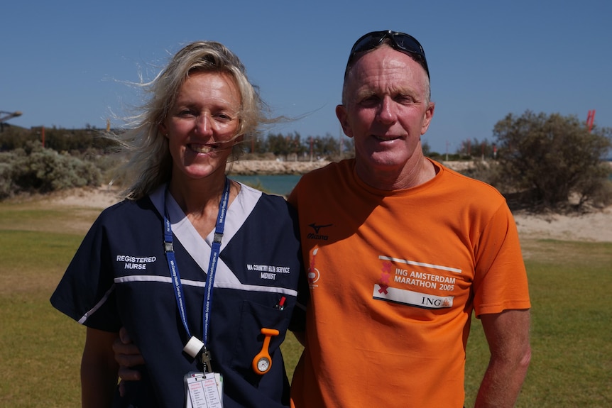 A woman in a blue nurse uniform and a man in an orange tshirt stand in front of a beach on a sunny day. 