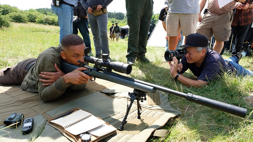 Will Smith (left) looks into sniper rifle scope as Ang Lee (right) looks through camera lens at him, both lying down on grass.