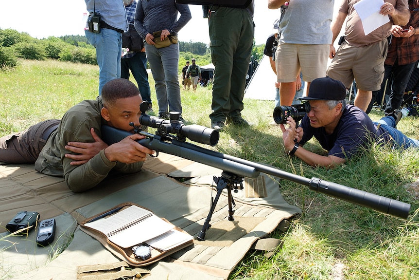 Will Smith (left) looks into sniper rifle scope as Ang Lee (right) looks through camera lens at him, both lying down on grass.