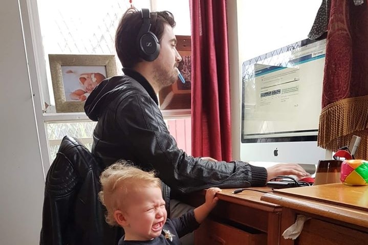A man wearing a headphone sits at a desk working, with a toddler crying and holding the desk next to him.
