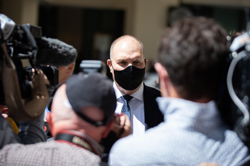 Man in mask stands in front of several cameramen and journalists.