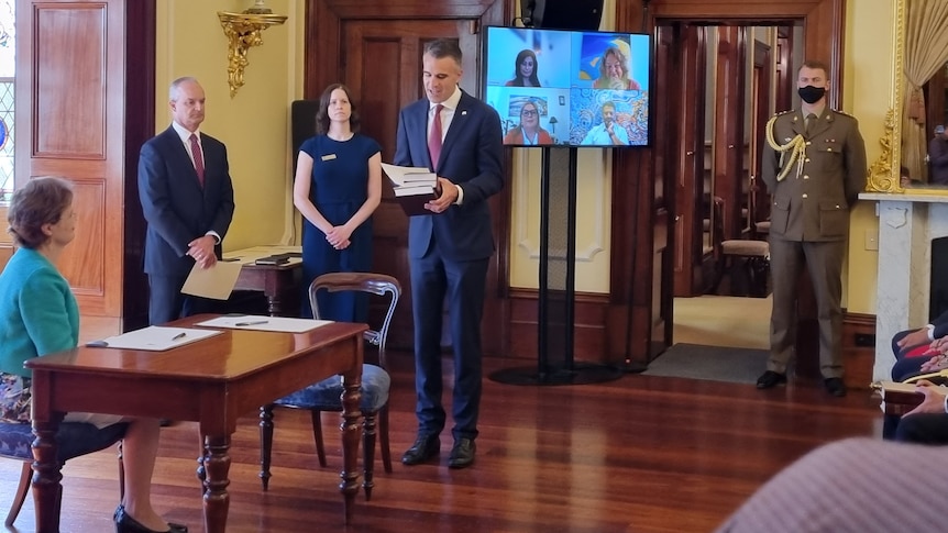 A man holds three books in front of a TV screen with people on it