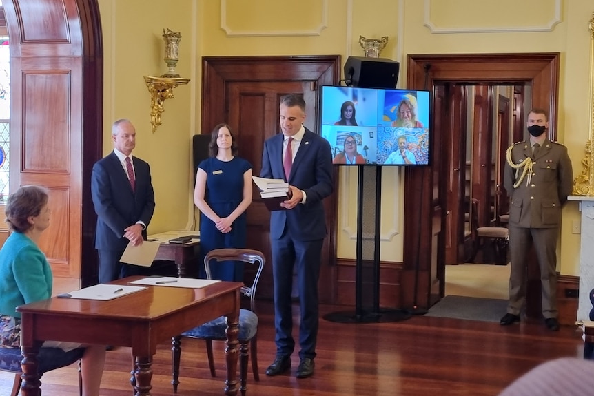 A man holds three books in front of a TV screen with people on it