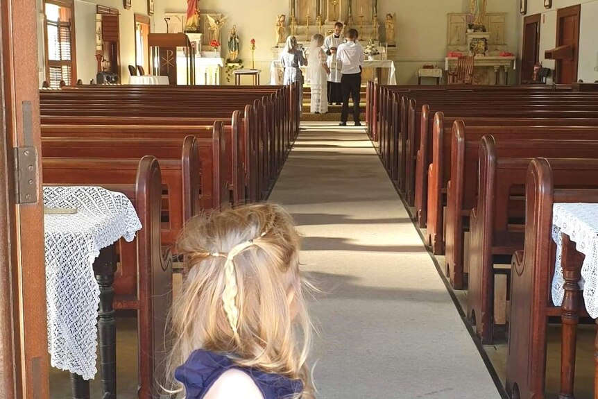 A young girl stares down the aisle of an empty church at a couple getting married at the altar.