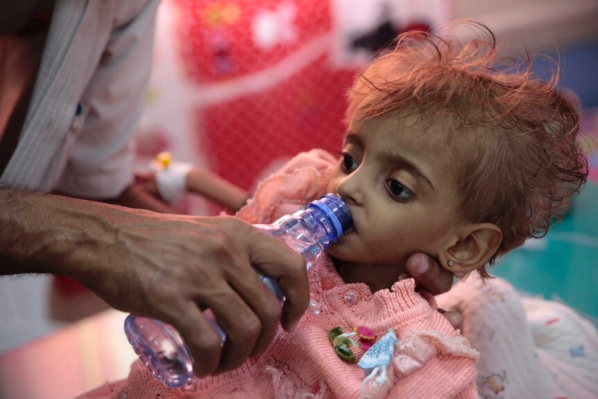 A malnourished Yemeni child wearing a pink knitted cardigan is given water through a bottle from her father.