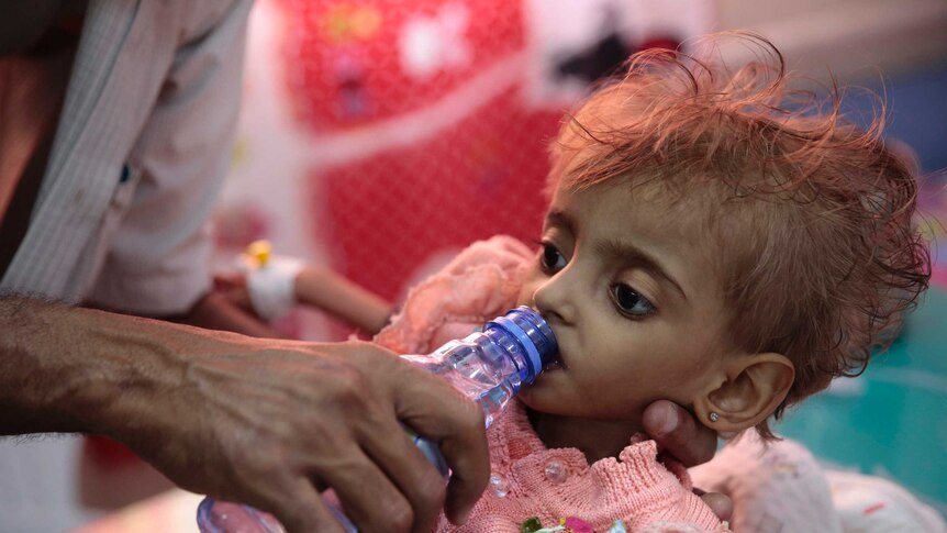 A malnourished Yemeni child wearing a pink knitted cardigan is given water through a bottle from her father.