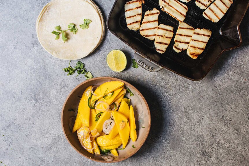 A stack of soft tacos, fried haloumi on a griddle and sliced mango and onion in a bowl in preparation to make taco recipe.