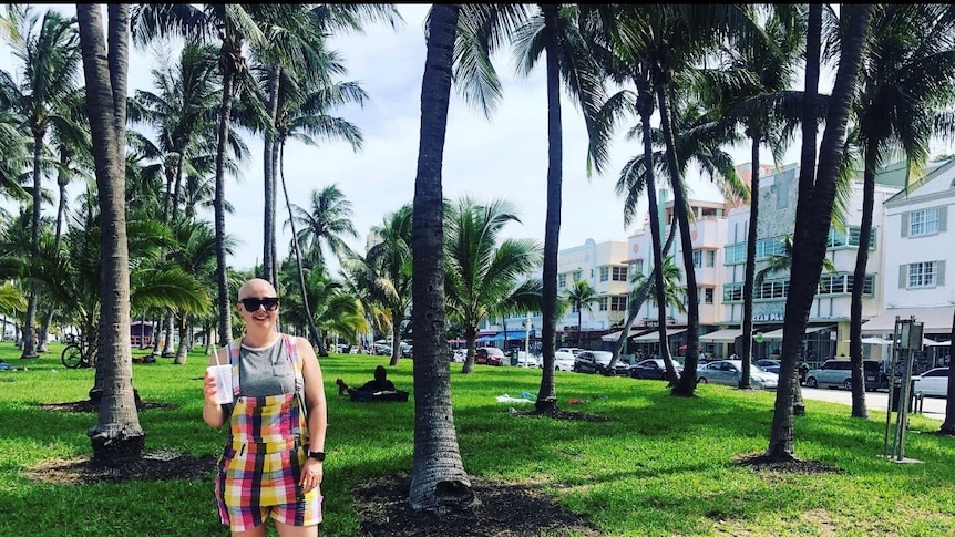 Woman with bald head stands in front of palm trees on holiday