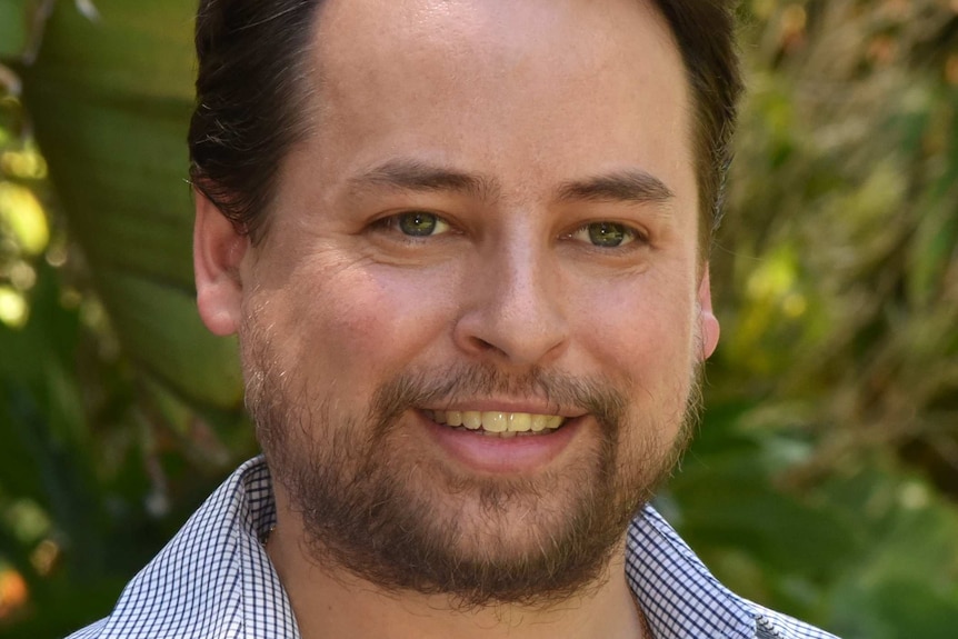 A man with brown hair smiles. He is wearing a blue and white cheque shirt with a collar. He has facial hair.
